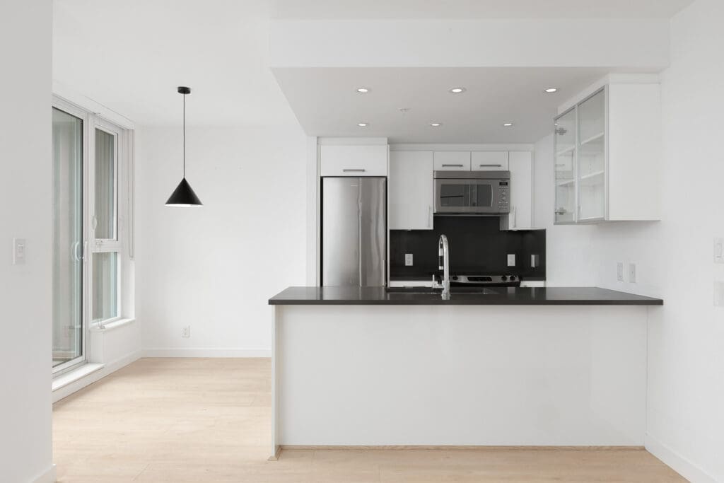 Modern kitchen with white cabinets, stainless steel appliances, black countertops, pendant light, and large window; open layout with light wood flooring.