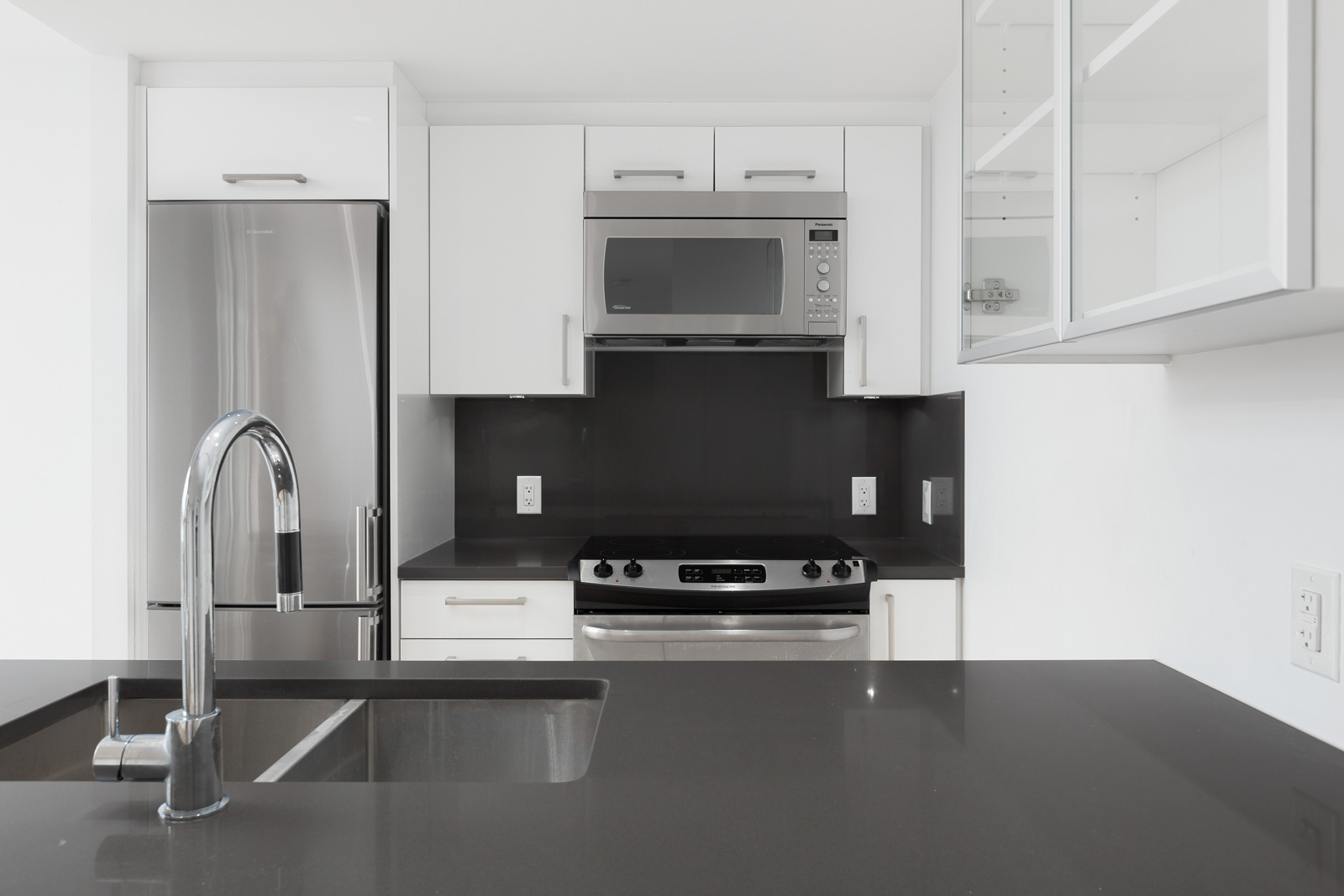 Modern kitchen with stainless steel appliances, black countertops, a built-in microwave, oven, refrigerator, and a sink with a faucet in the foreground.