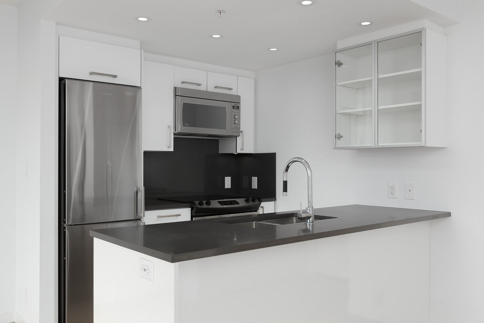 Modern kitchen with stainless steel appliances, black countertops, white cabinets, and a sink on a central island. The upper right cabinet doors are open, showing empty shelves.