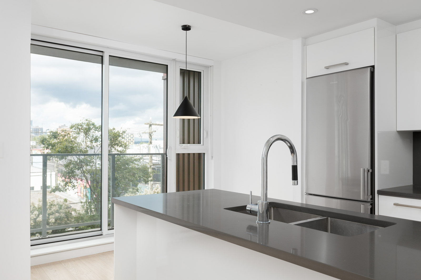 A modern kitchen with a grey countertop island, built-in sink, stainless steel fridge, pendant light, and large sliding glass doors leading to a balcony.