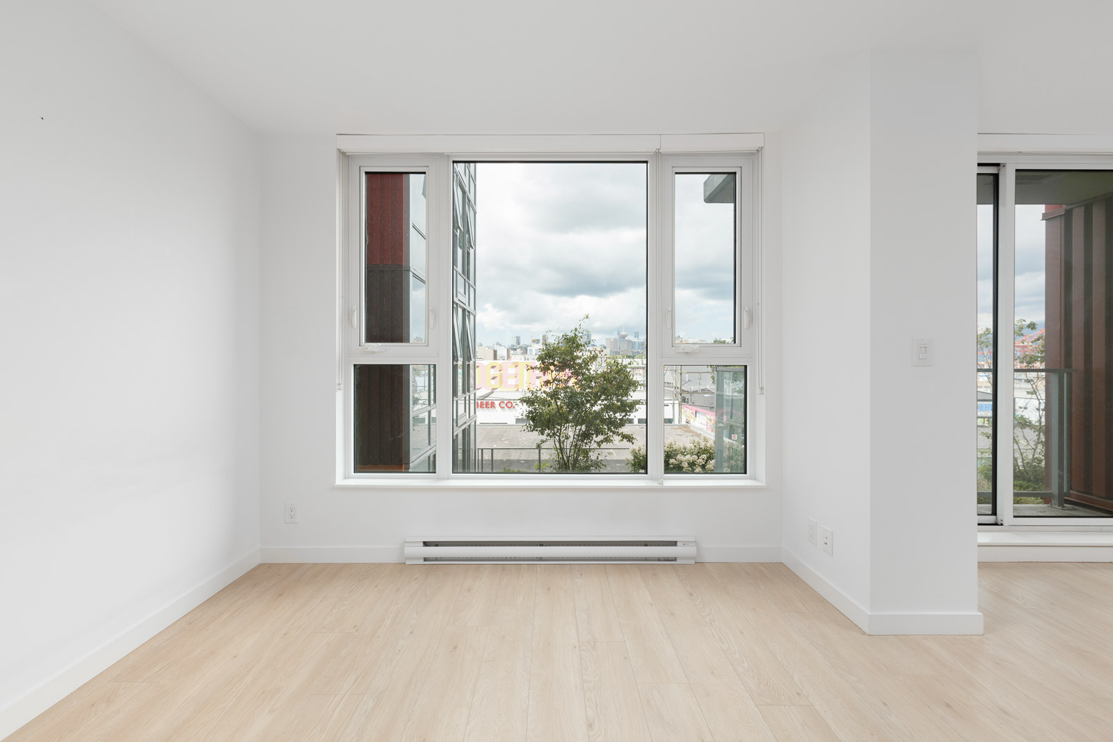 Empty room with light wood flooring, white walls, and large windows overlooking a cityscape with cloudy sky.