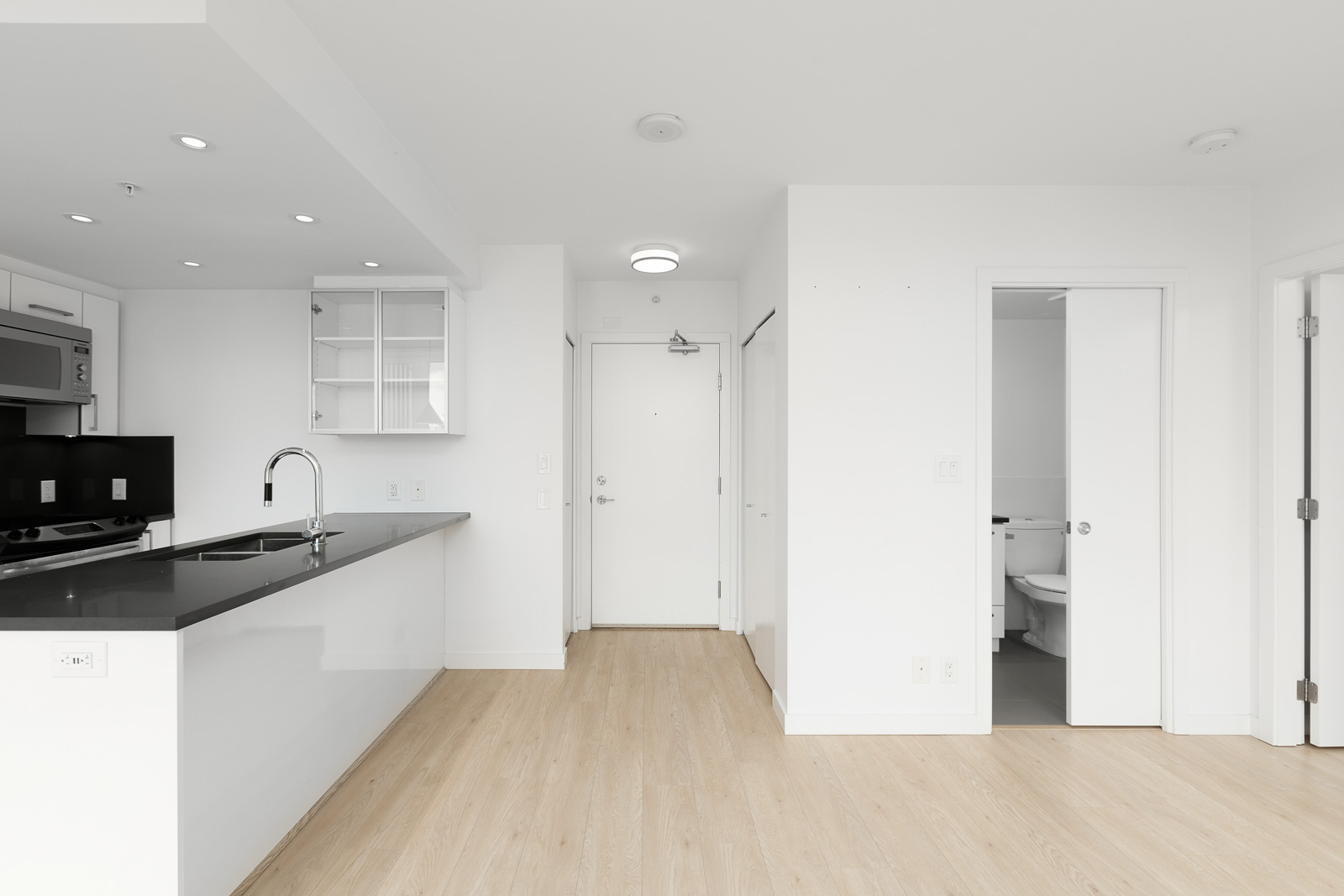 Modern, unfurnished apartment interior with light wood floors, white walls, a kitchen area on the left, a closed entrance door in the center, and a bathroom visible on the right.