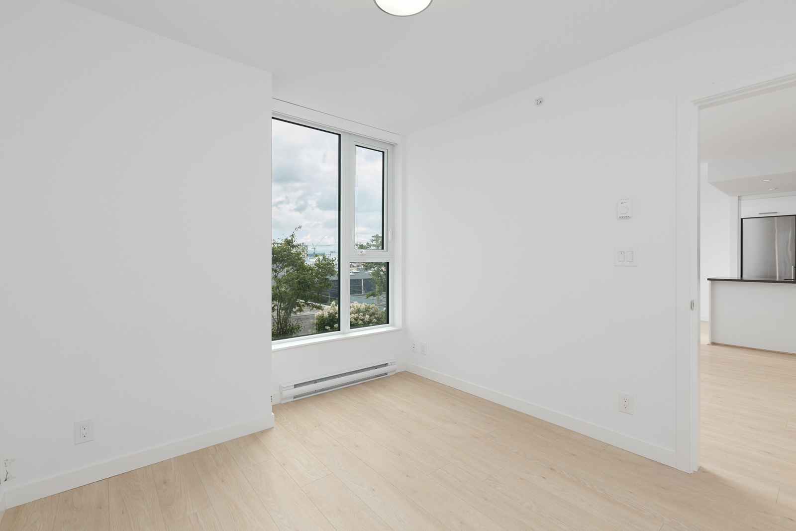 Unfurnished, empty white room with a large window, light wood floors, and an open doorway leading to a modern kitchen with stainless steel appliances.