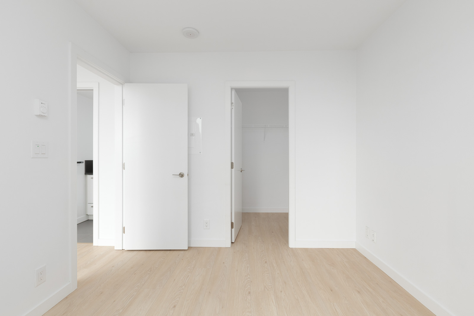 Empty white room with light wood flooring, two open doors—one leads to a closet, the other to another room. Walls and ceiling are plain and white.