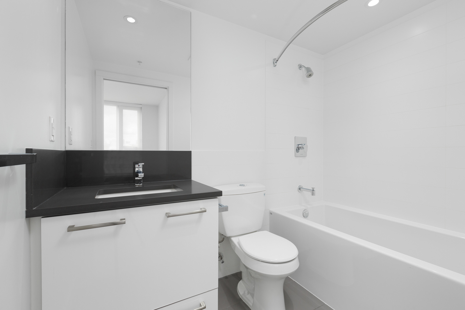 Modern white bathroom with a black countertop, sink, toilet, bathtub, wall-mounted shower, and large mirror above the sink.
