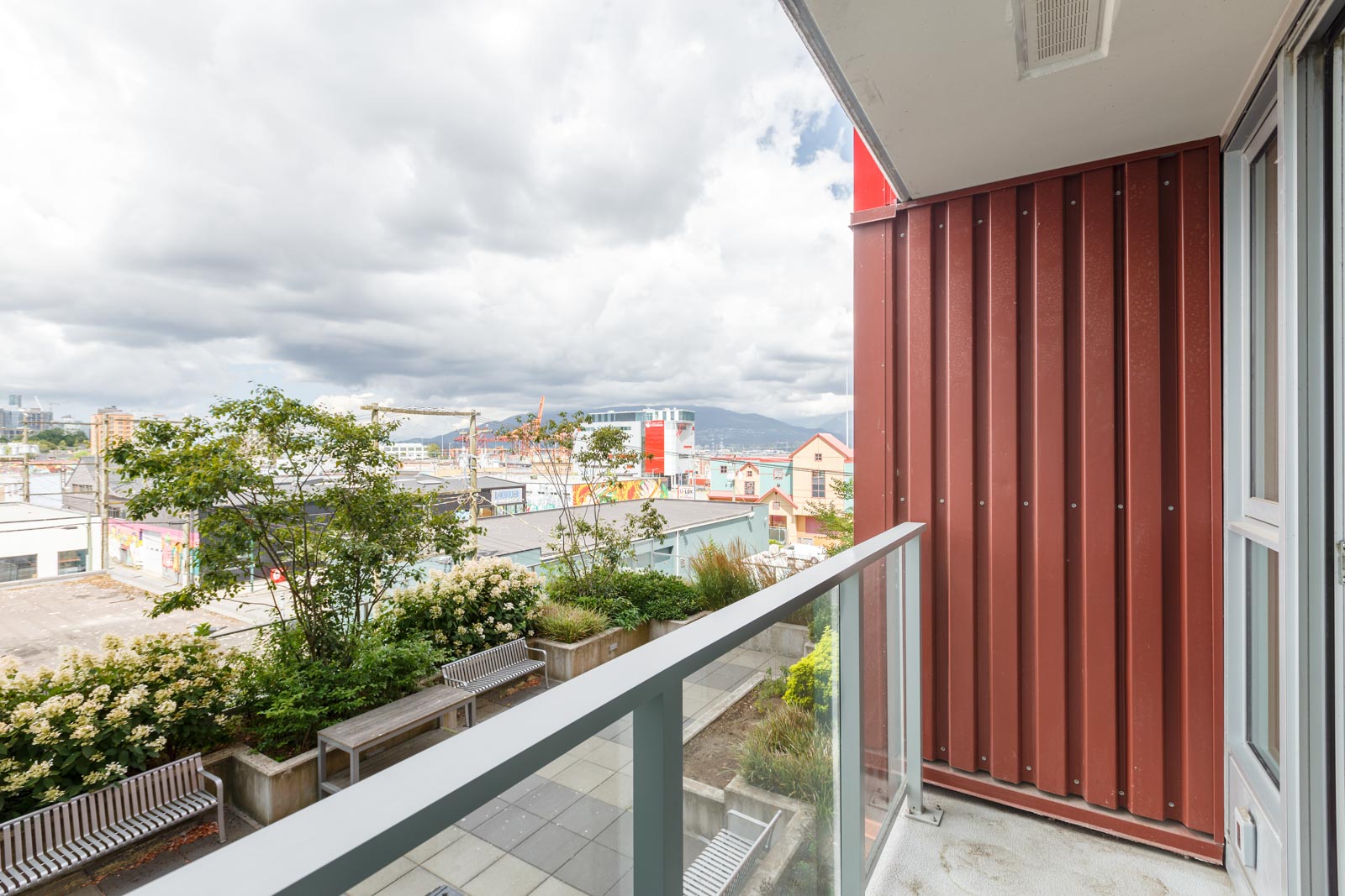 View from a balcony with glass railing, overlooking an urban street with buildings, plants, benches, and a cloudy sky.