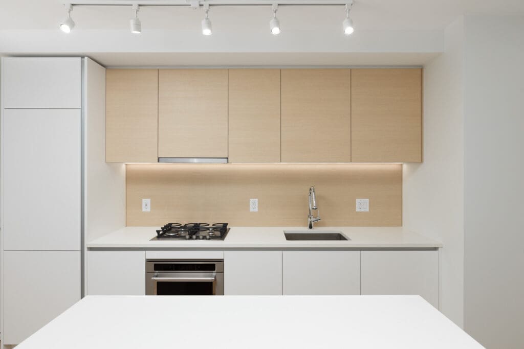 Minimalist kitchen with light wood cabinets, white countertops, a gas stove, built-in oven, sink with faucet, and ceiling track lighting.