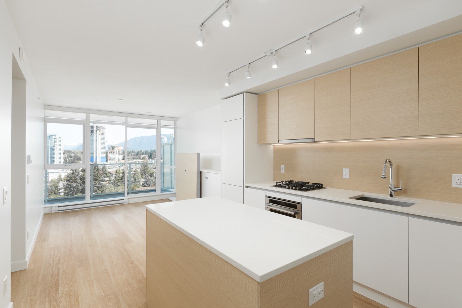Modern kitchen with light wood cabinets, white countertops, an island, built-in appliances, and large windows with a city and mountain view in the background.