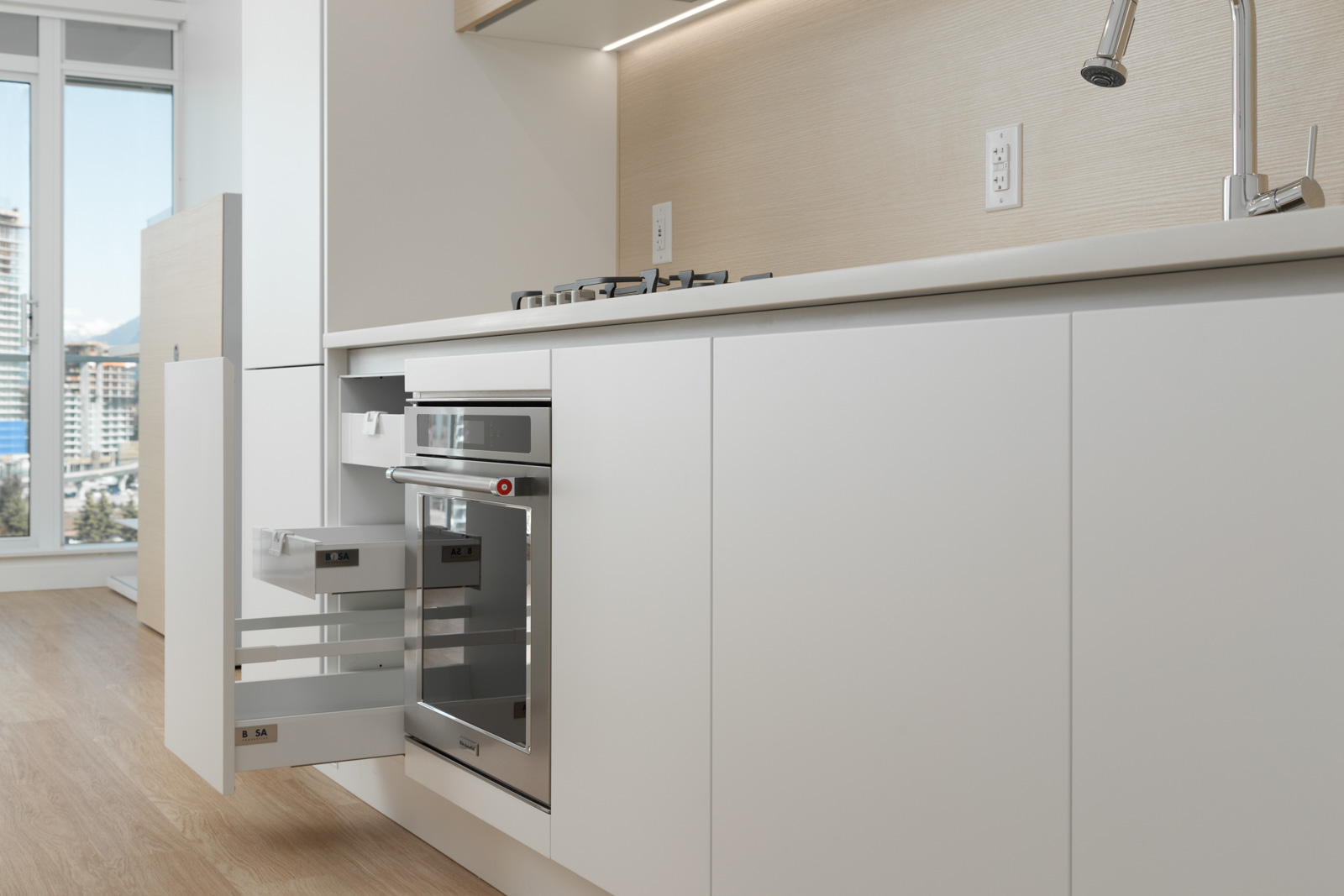 Modern kitchen with white cabinets, open drawers, a built-in oven, gas stove, and a window showing a cityscape in the background.