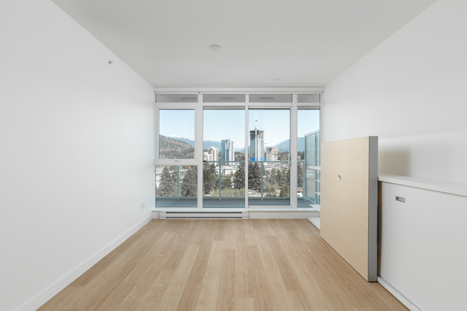Unfurnished room with light wood flooring, white walls, large windows, cityscape and mountains visible outside, and a fold-down bed against the wall.