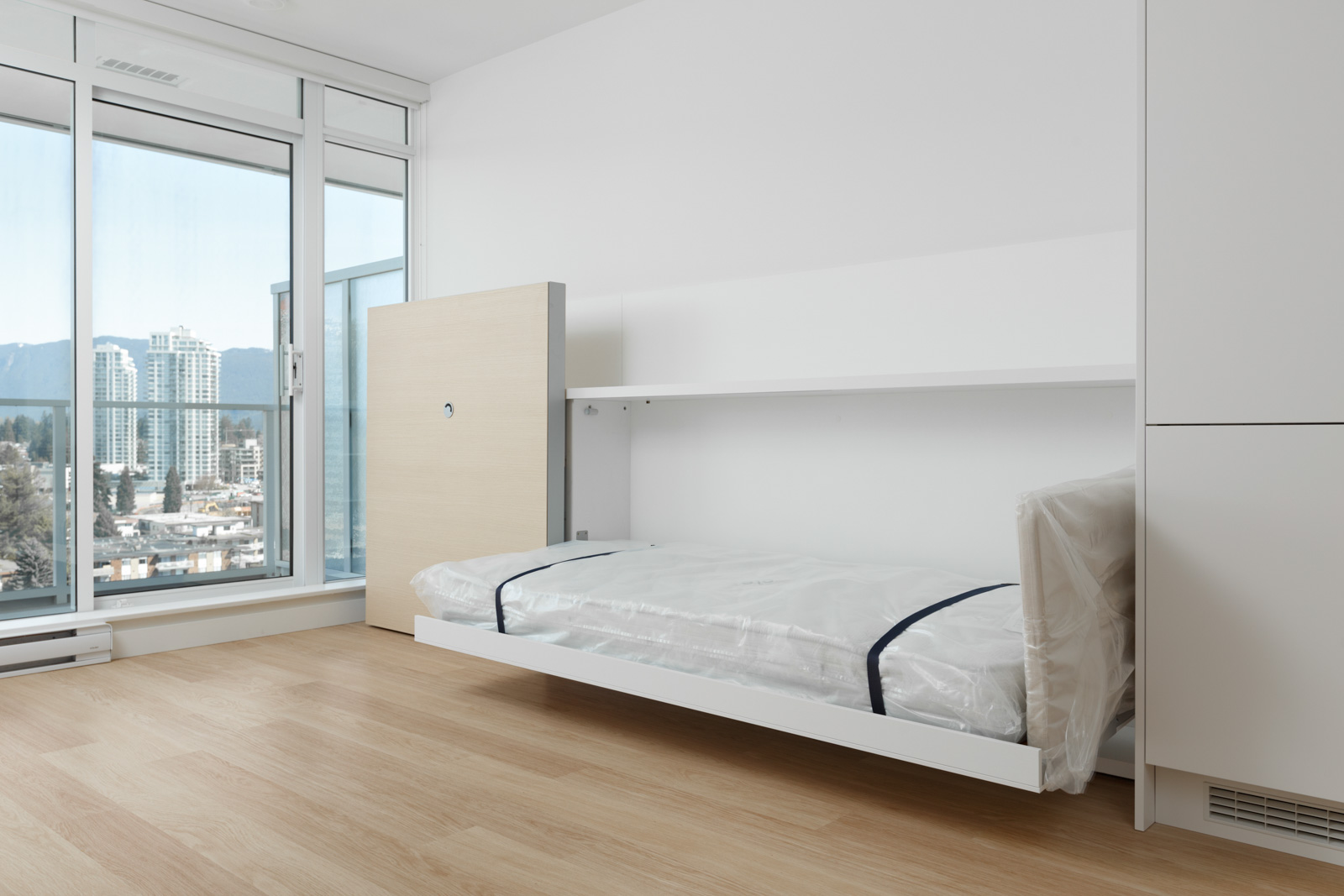 A modern, unfurnished room with a Murphy bed partially folded down, large windows, and a city view in the background.