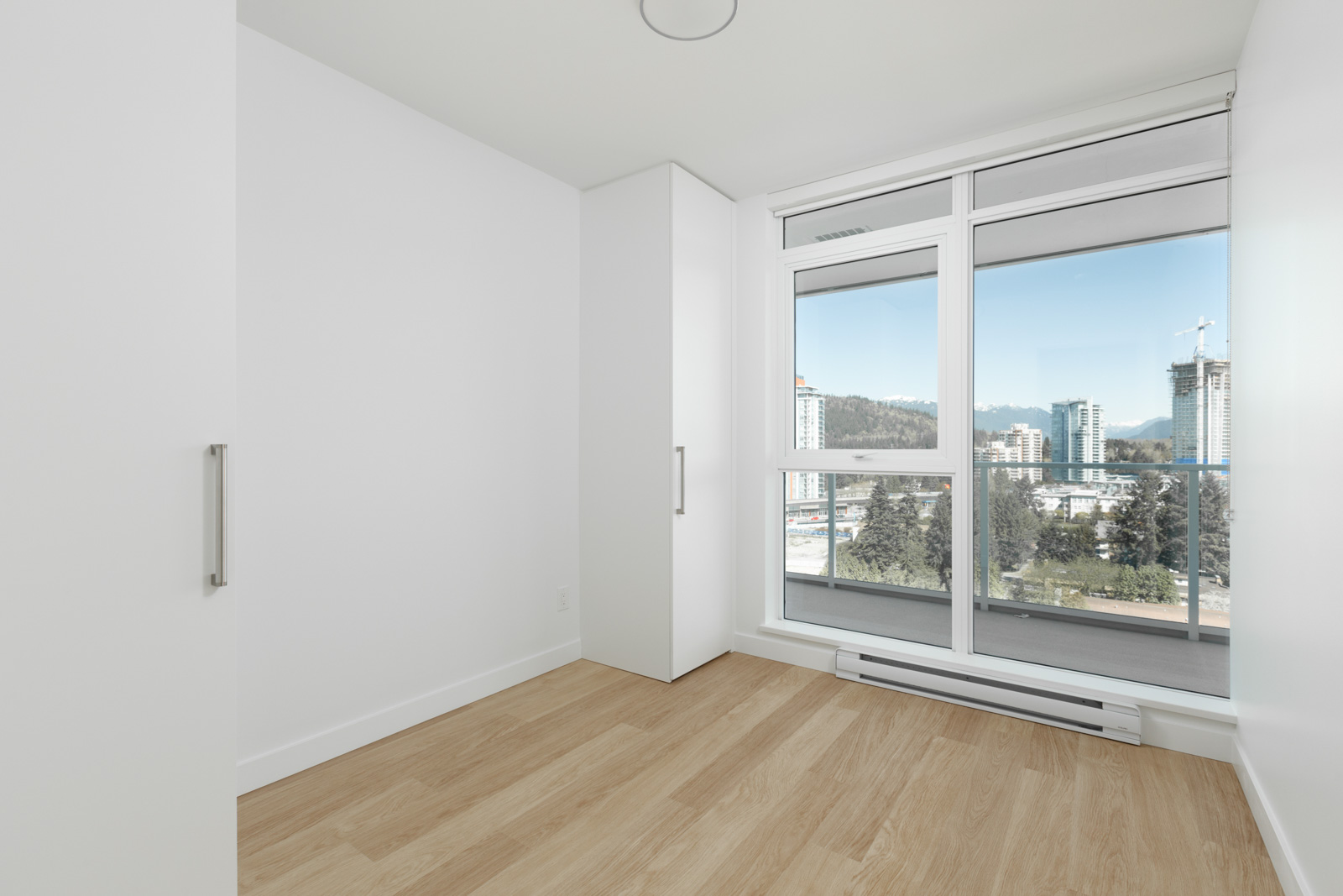 Empty room with light wood flooring, white walls, built-in closet, and large window showing a cityscape with tall buildings and trees outside.