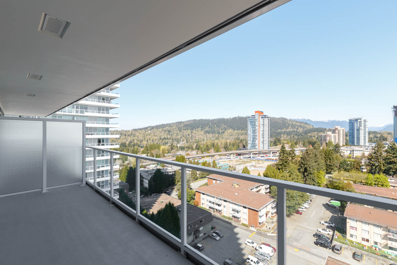 High-rise balcony with glass railing overlooking residential buildings, roads, and distant forested hills under a clear sky.