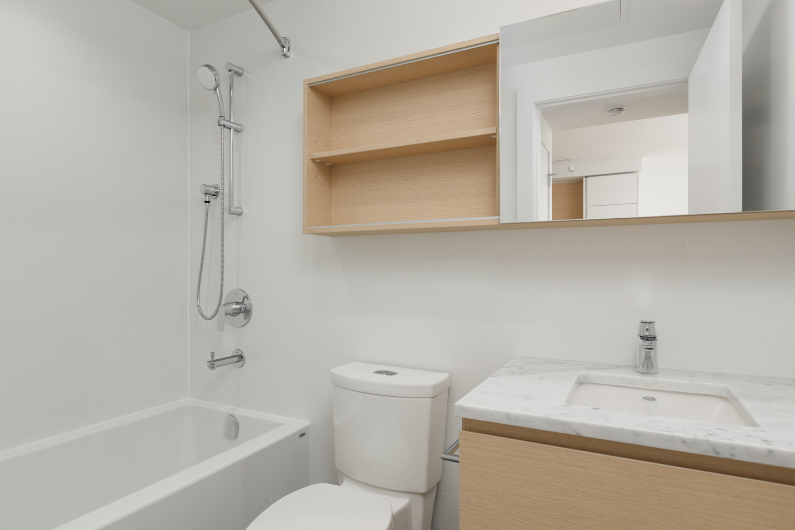 Modern bathroom with a bathtub-shower combo, toilet, light wood cabinet with a sink, wall mirror, and open shelving above the toilet.