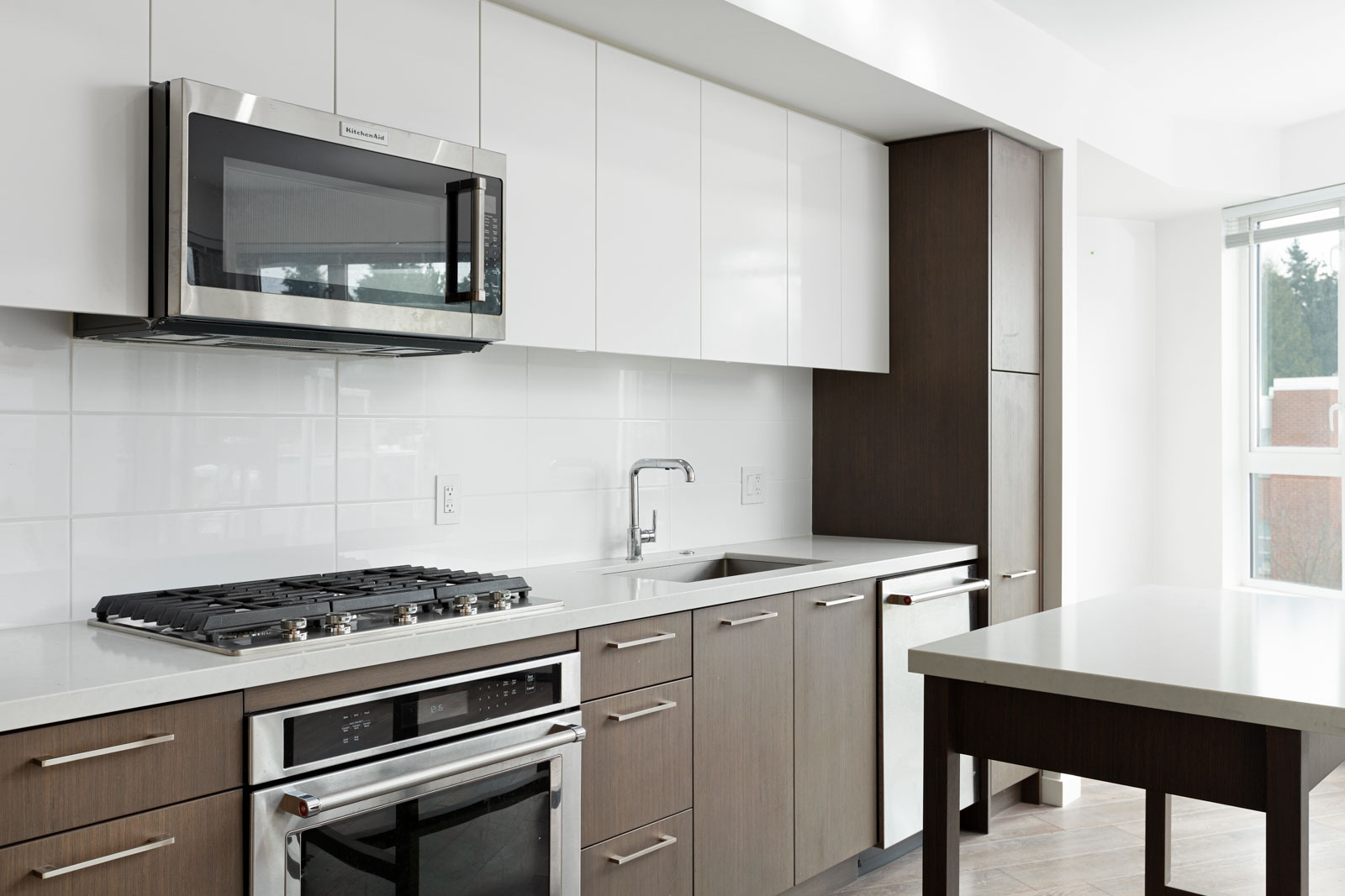 Modern kitchen with stainless steel appliances, gas stove, built-in microwave, white cabinets, brown lower cabinets, and large window providing natural light.