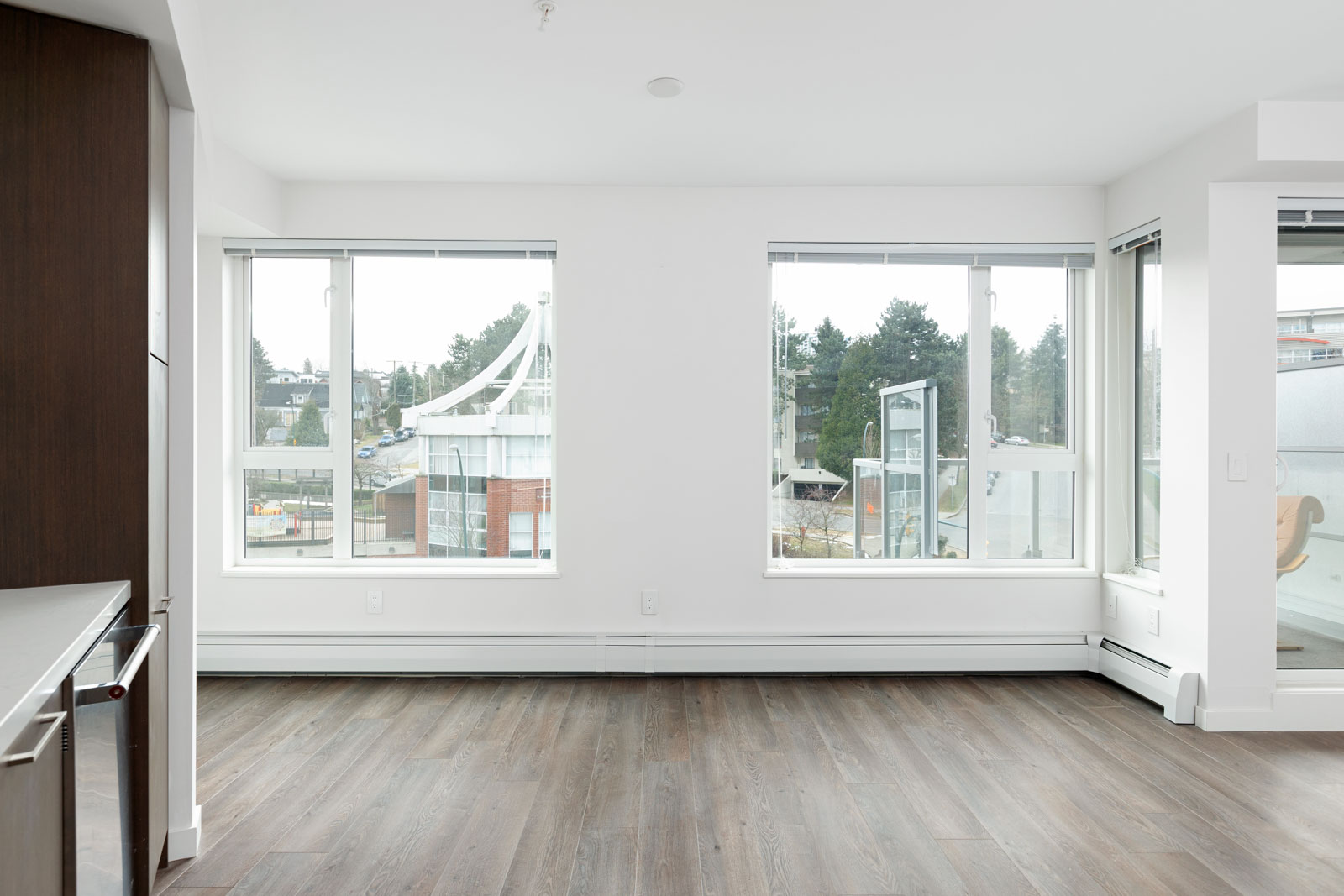 Bright, empty room with large windows, wood flooring, and white walls, overlooking an urban area with trees and buildings outside.