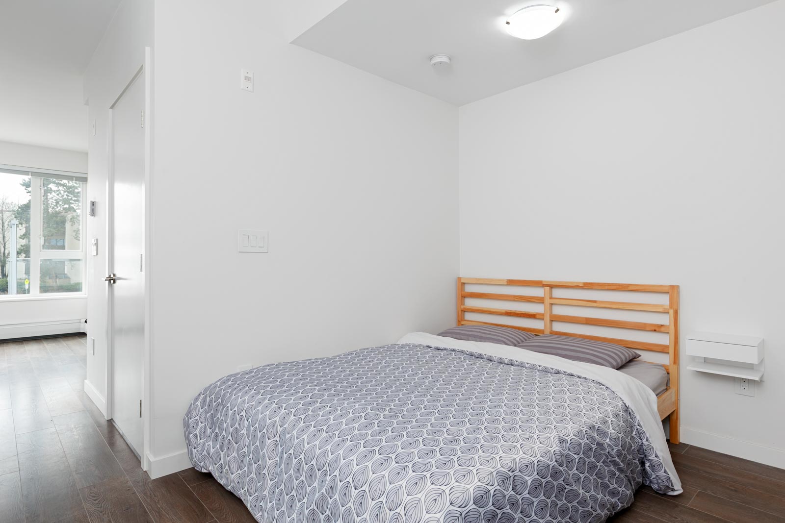 A minimal bedroom with a wooden bed frame, gray patterned bedding, white walls, and dark wood flooring. A window and door are visible in the background.