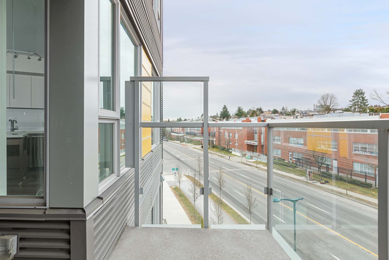 A modern apartment balcony with glass railing overlooks a quiet urban street and commercial buildings on a cloudy day.