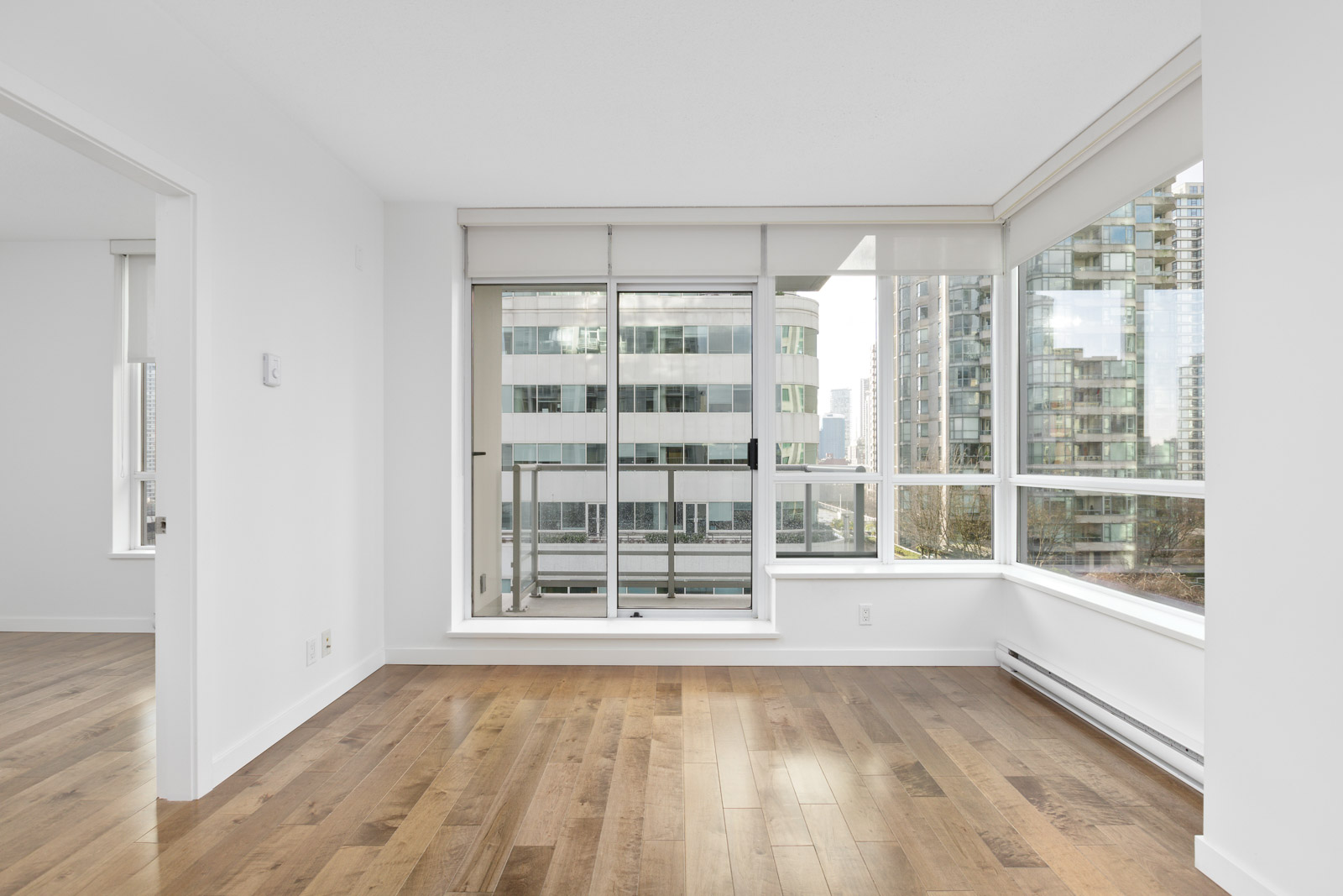 Unfurnished modern apartment room with large windows, wooden floor, and city buildings visible outside. Adjacent room partially visible on the left.