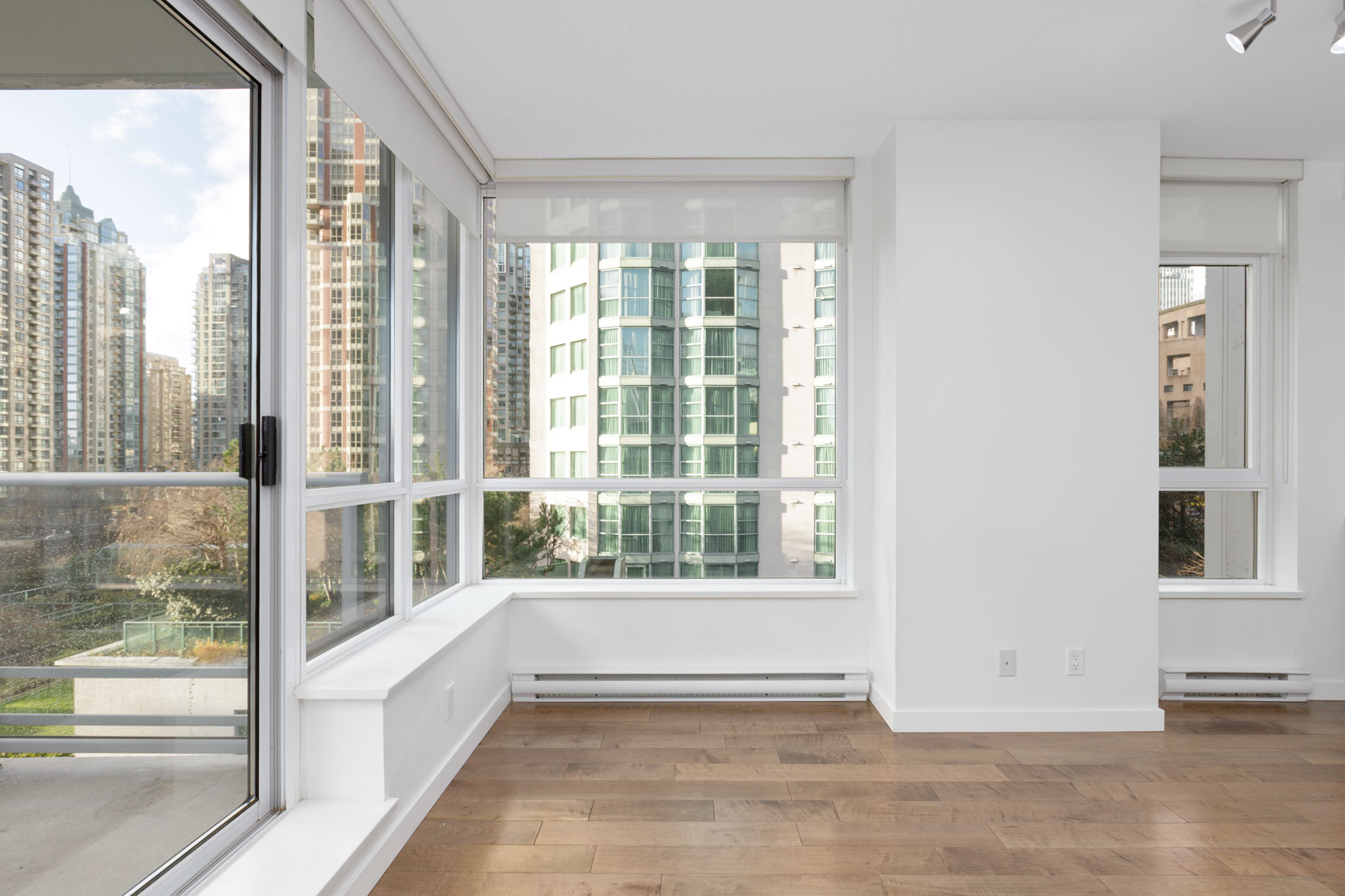 Empty modern apartment with large windows, white walls, hardwood floors, and city buildings visible outside.