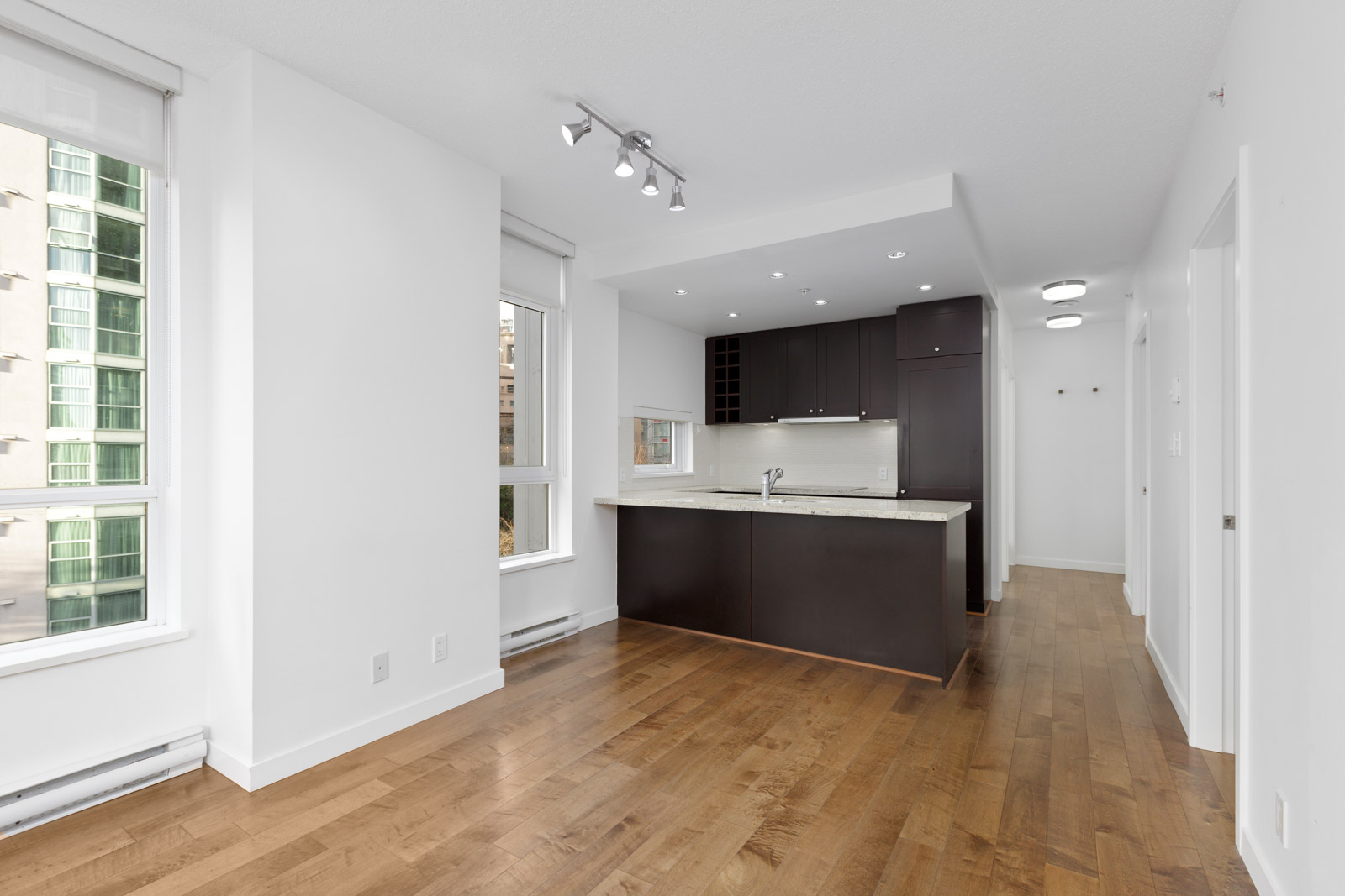 Modern apartment interior with hardwood floors, white walls, large windows, and an open kitchen featuring dark cabinets and a countertop.