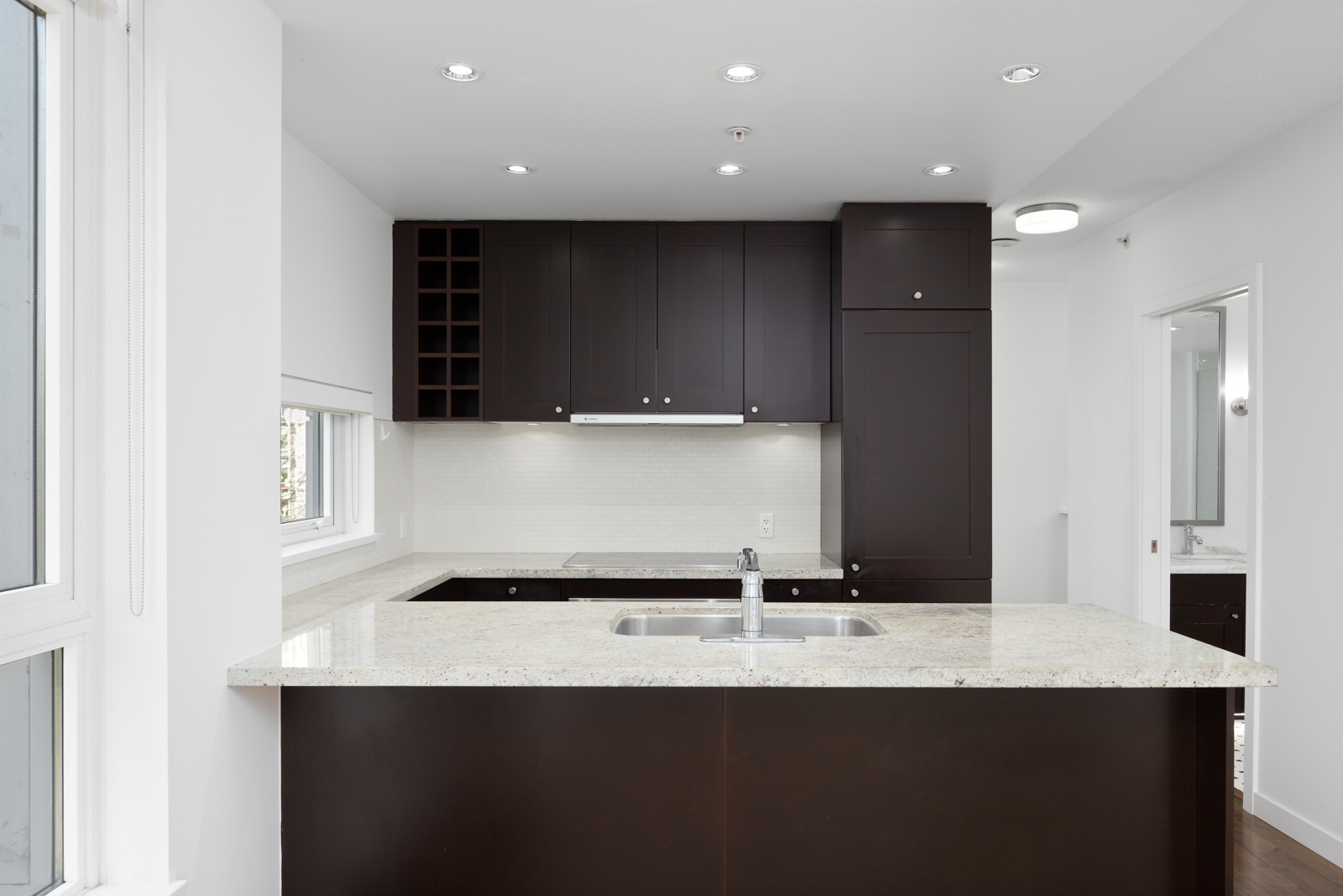 Modern kitchen with dark cabinets, white countertops, built-in sink, and overhead lighting. Large windows provide natural light.