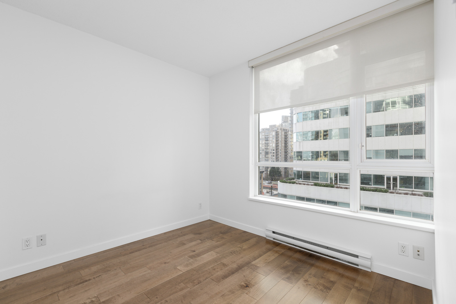 Empty room with white walls, light wood floor, large window with a roller shade, and a view of modern city buildings outside.
