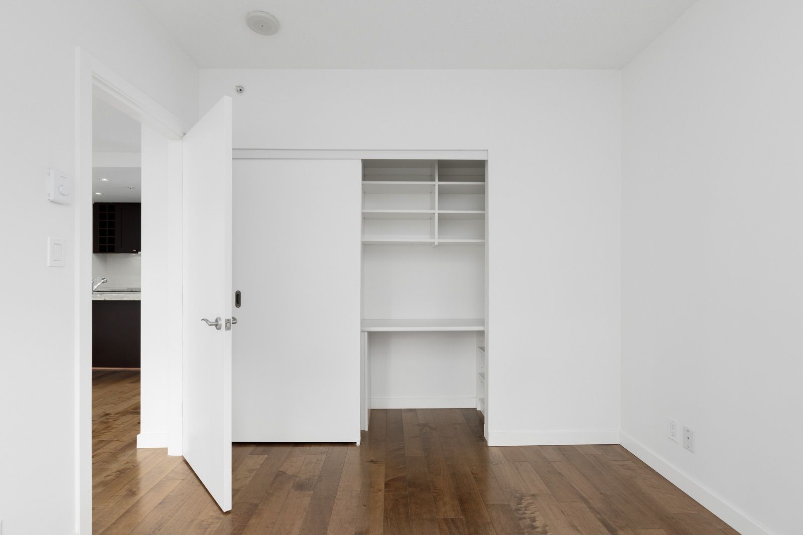 Empty white room with wooden floor, open closet with shelves, and open door leading to a kitchen area.