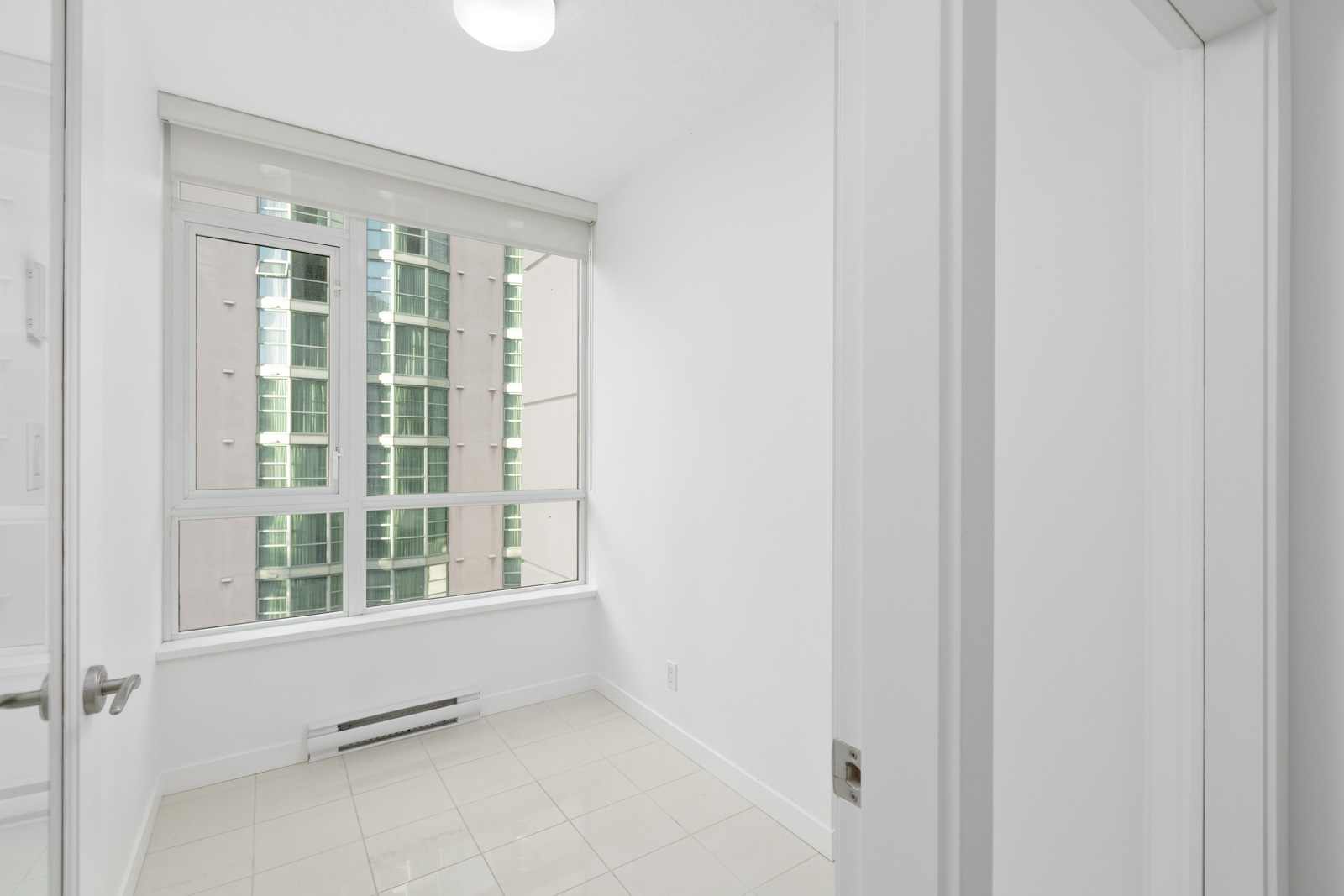A small, empty white room with tiled floor, large window, and view of neighboring building. Door is partially open in the foreground.