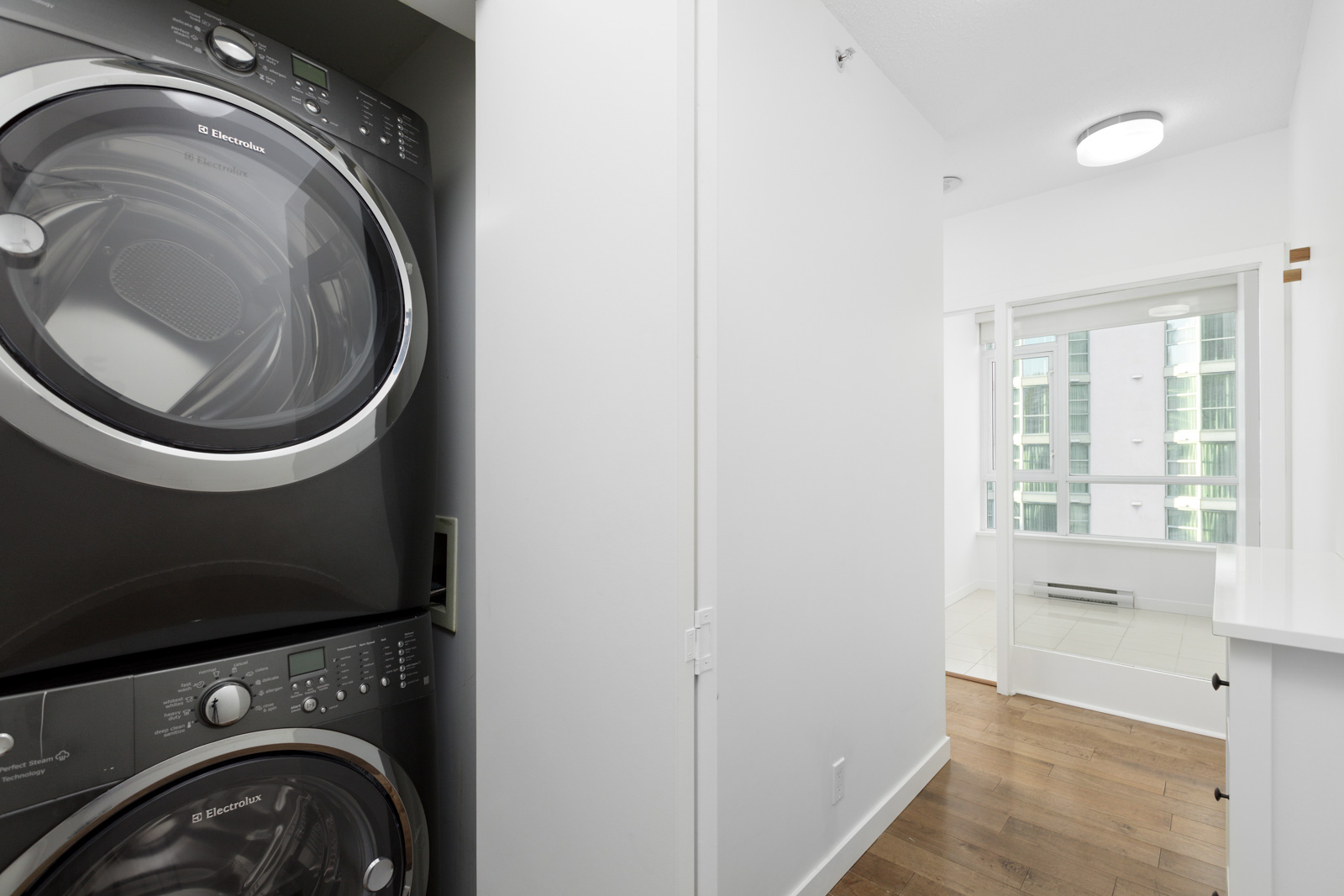 Stacked black washer and dryer in a narrow laundry area with white walls, wood floors, and a view of another room with large windows.