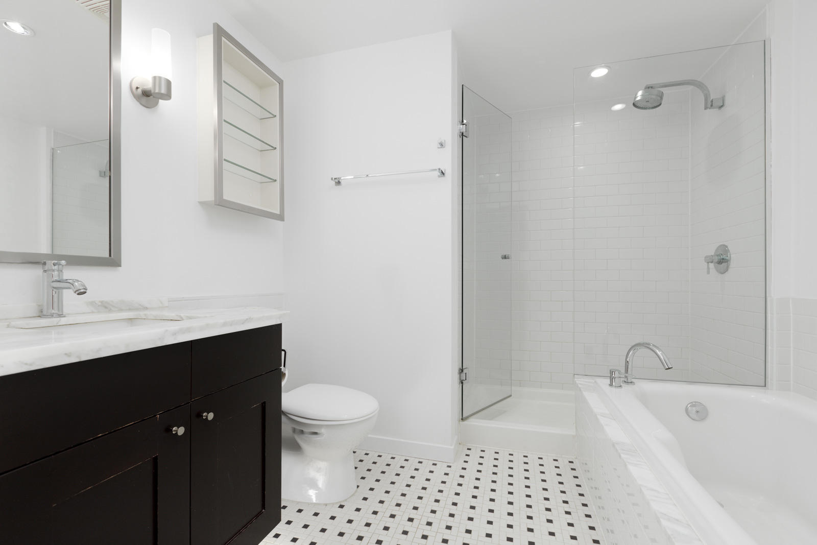 Modern bathroom with black vanity, white countertop, toilet, glass shower, built-in shelves, bathtub, and white tile flooring with black accent tiles.