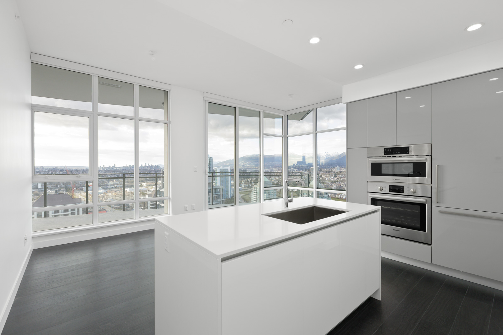 Modern kitchen with white island, built-in stainless appliances, and floor-to-ceiling windows offering a city and mountain view.
