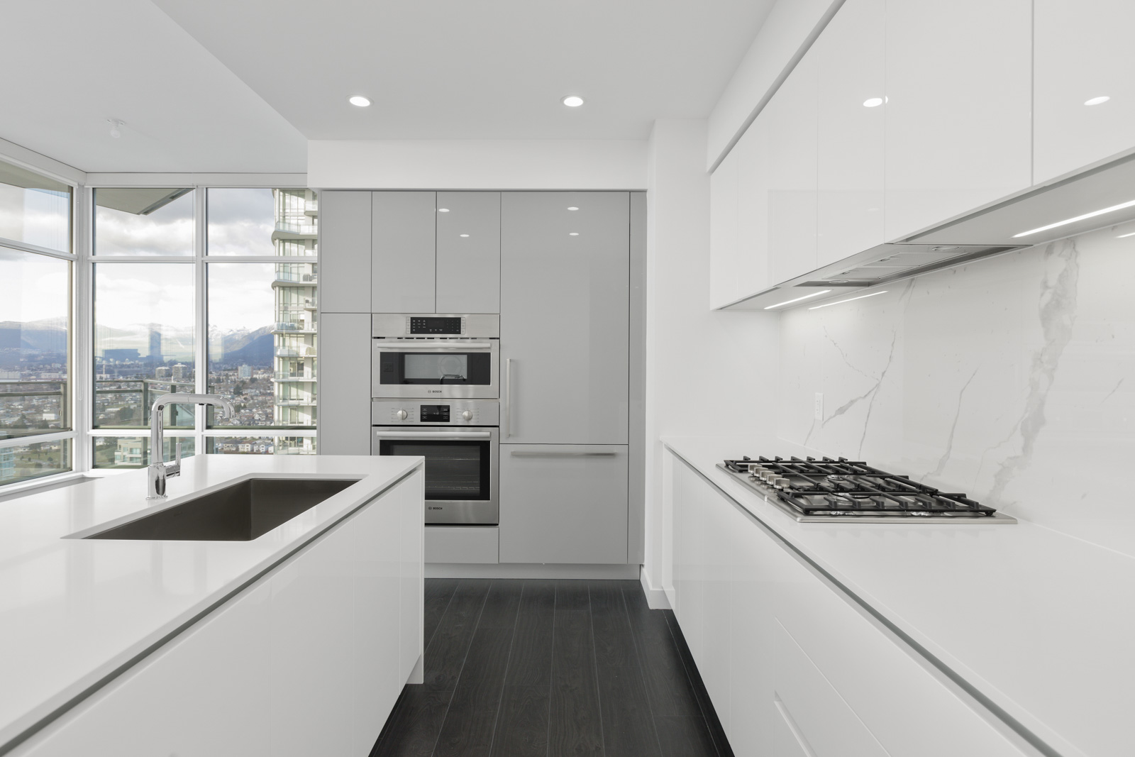 Modern kitchen with white cabinets, built-in stainless steel appliances, gas cooktop, marble backsplash, and large windows with a city view.