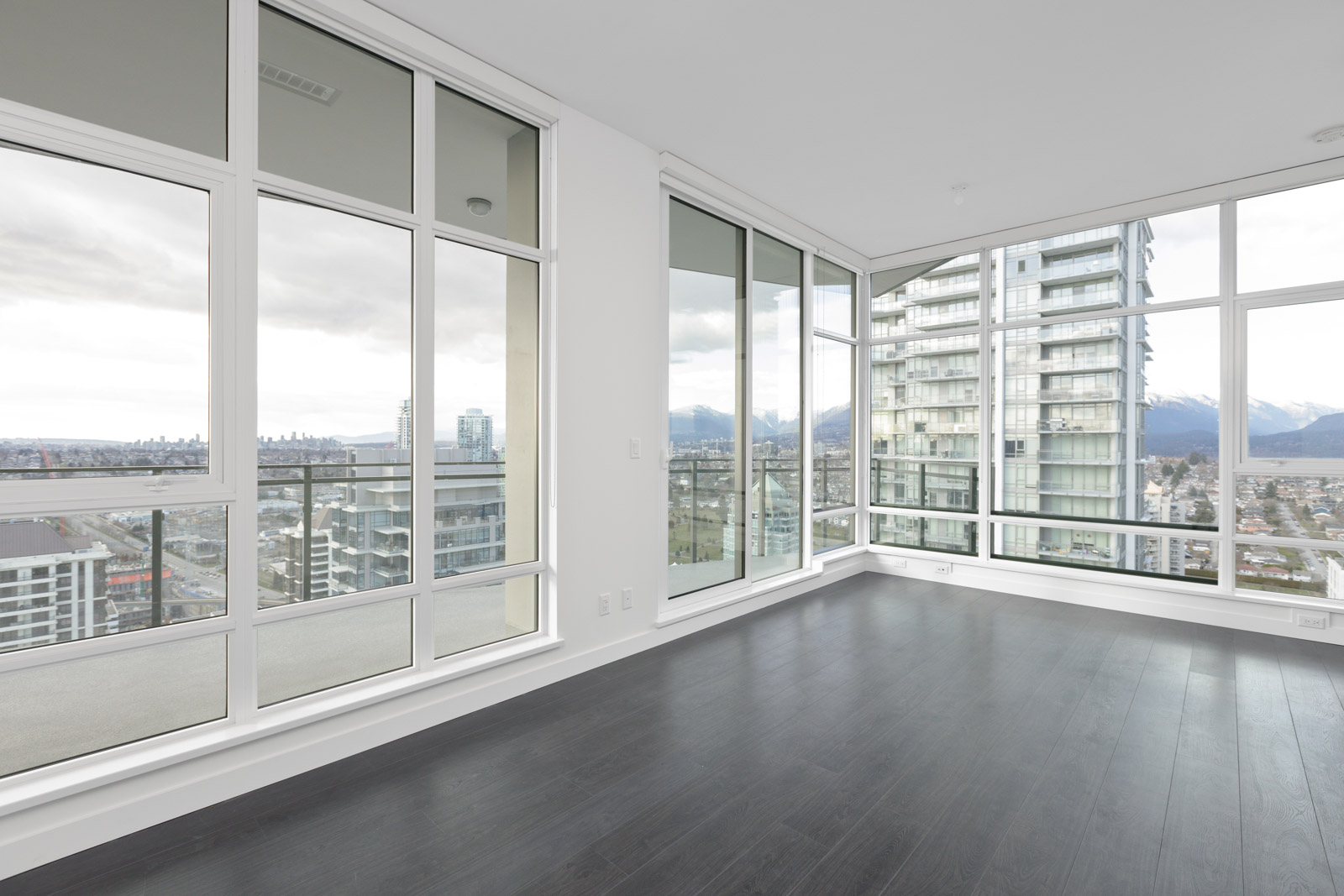 Empty modern apartment corner with floor-to-ceiling windows, dark wood flooring, and a view of city buildings and mountains outside.