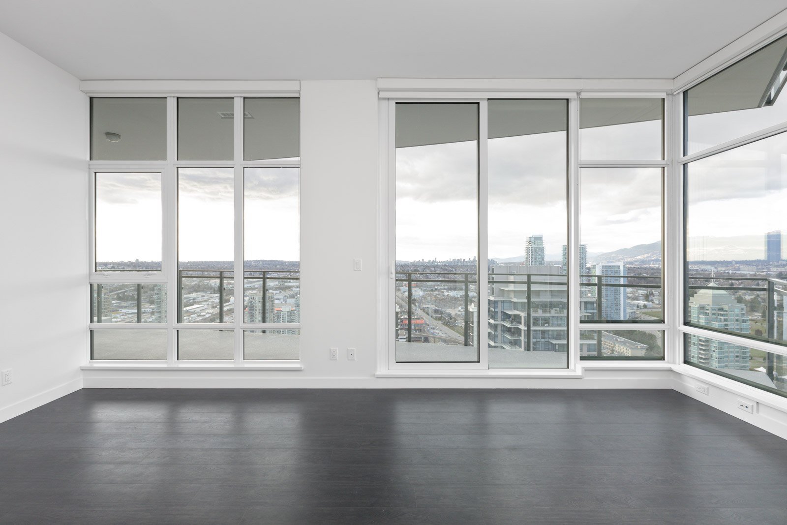 Modern, empty room with dark hardwood floors, large floor-to-ceiling windows, and a cityscape view under a cloudy sky.