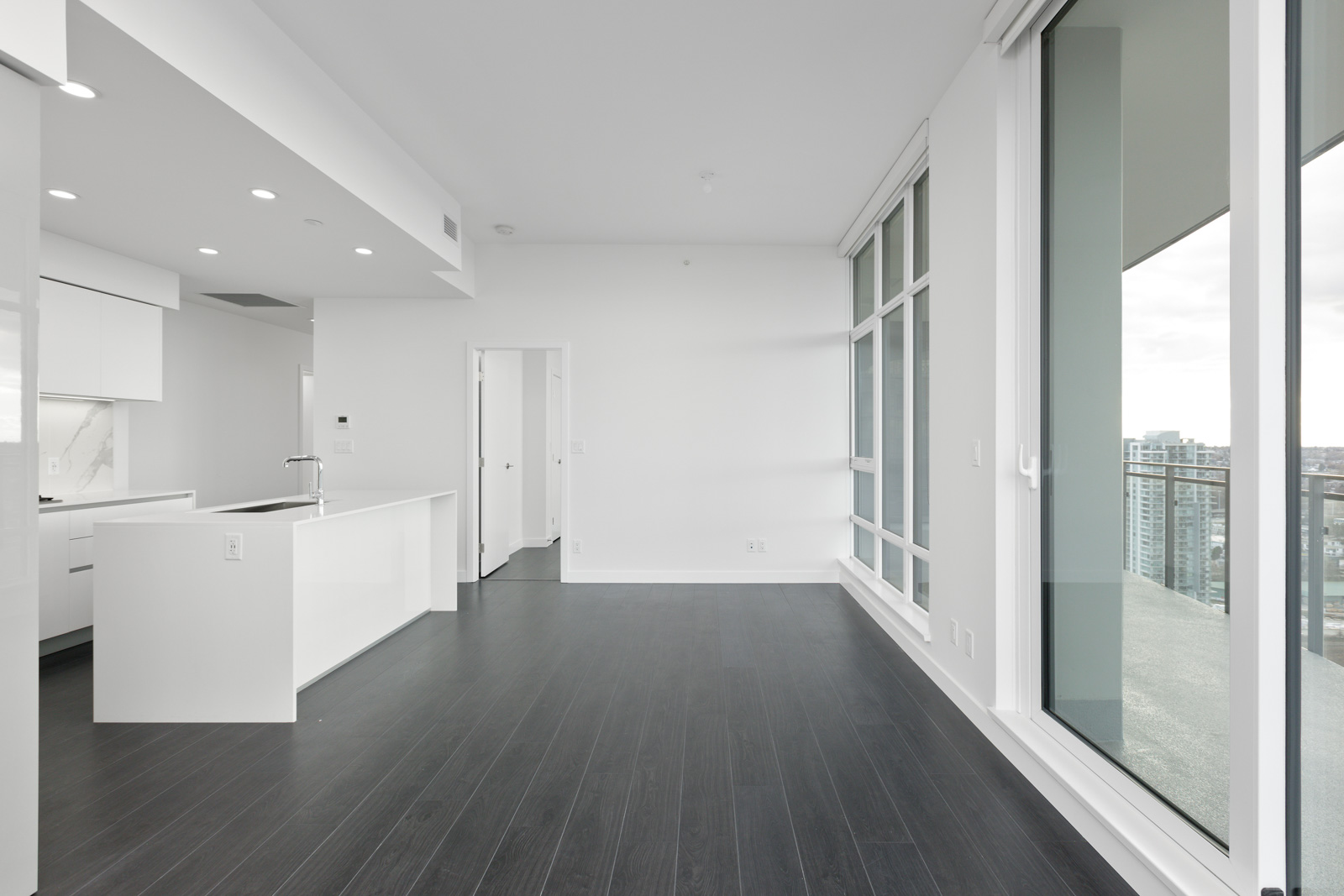 Modern empty apartment with dark wood floors, white walls, large windows, an open kitchen with an island, and a view of the city from a high-floor balcony.
