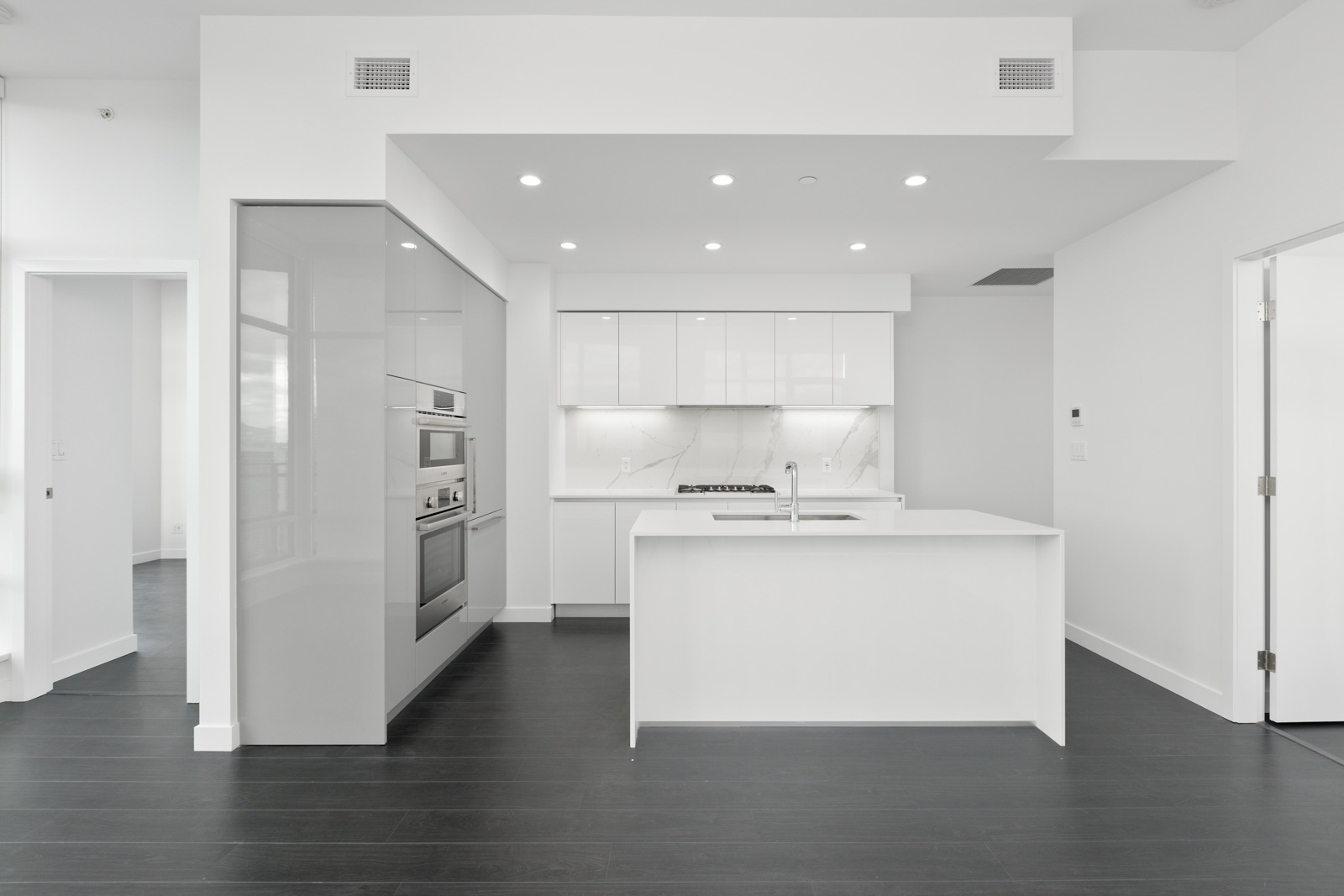 Modern white kitchen with an island, built-in appliances, glossy cabinets, and dark wood flooring. The kitchen opens to adjacent rooms with minimalist design.