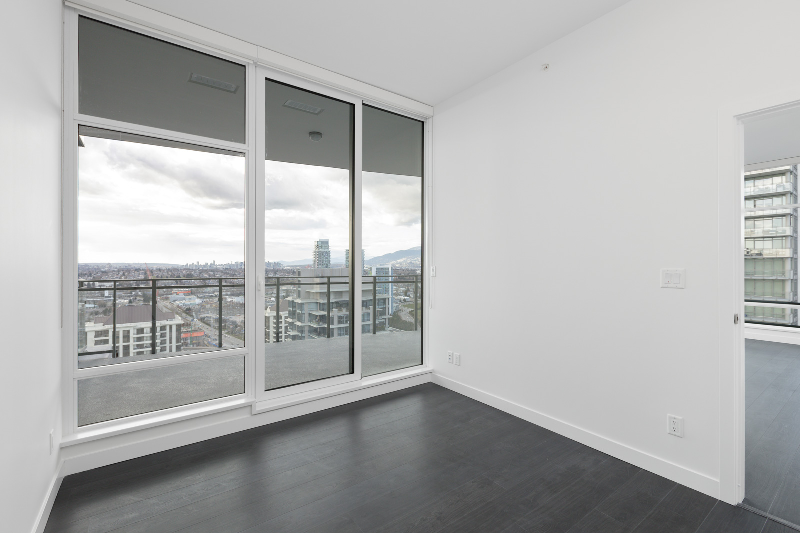 Unfurnished modern apartment room with dark floors, large glass doors leading to a balcony, and floor-to-ceiling windows showing a cityscape view.