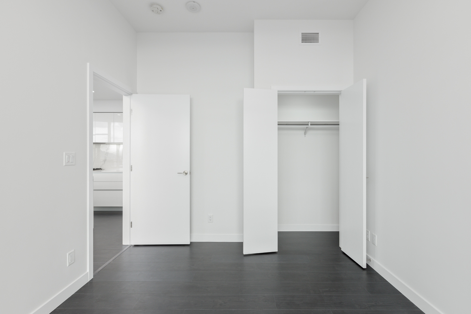 Empty white room with dark flooring, an open door leading to another room, and an open closet with double doors.