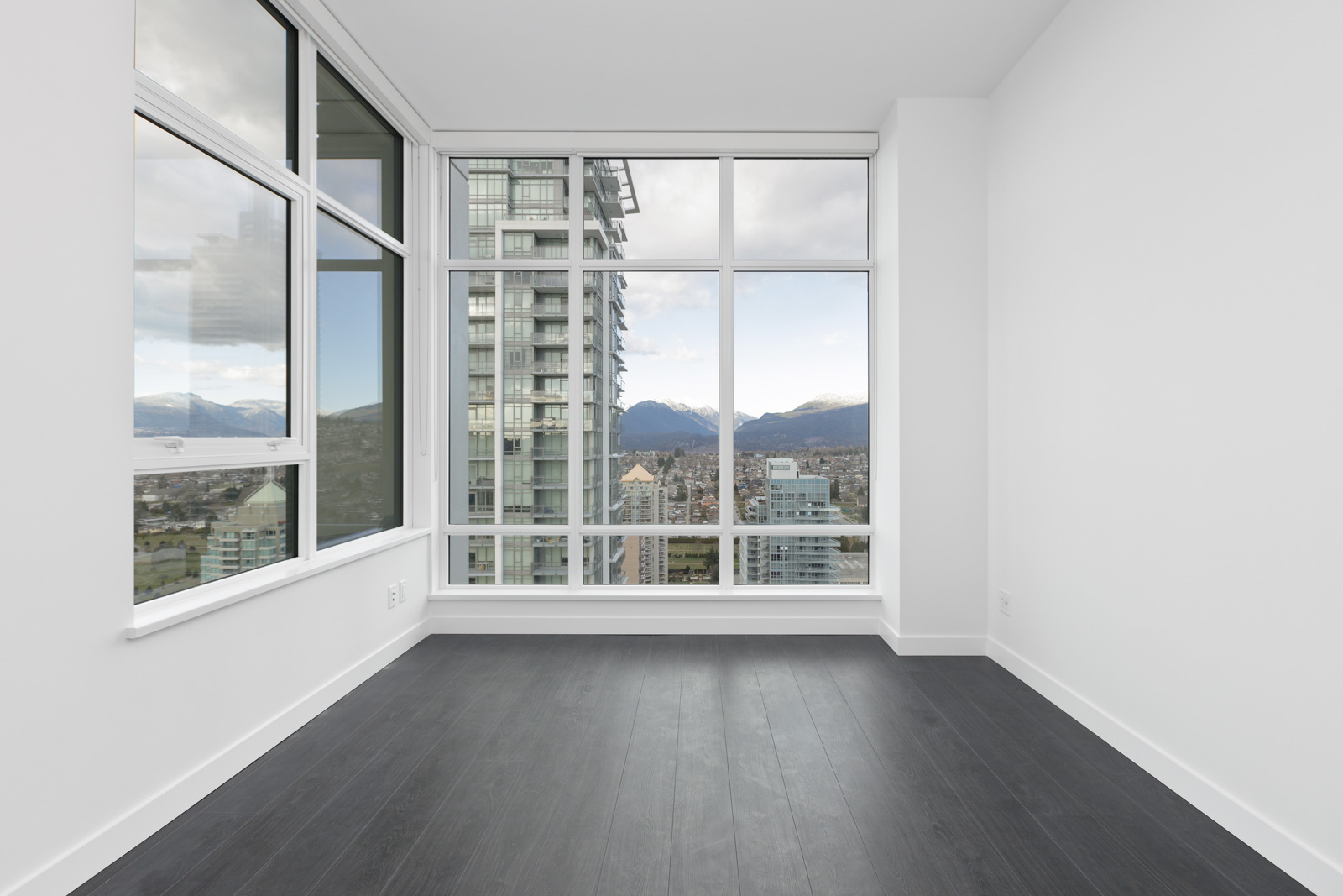 Empty modern room with large windows, dark wood floor, and a view of city buildings and mountains in the background.