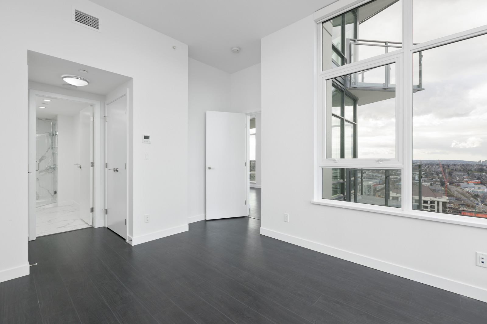 Modern empty room with dark wood floors, white walls, large windows showing a city view, and an open door leading to a hallway and bathroom.