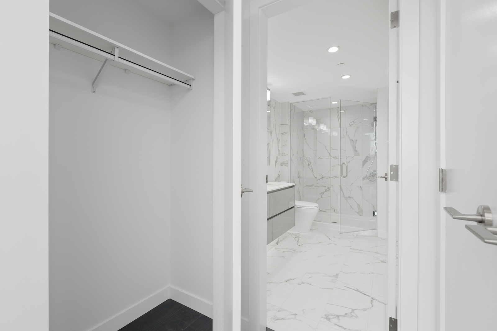 View into a modern bathroom with marble tile flooring and walk-in shower, seen from an adjacent empty white closet with a hanging rod and shelf.