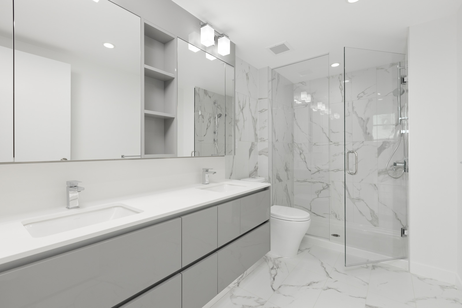 Modern bathroom with gray cabinets, double sinks, large mirror, white marble tile floors and walls, glass shower enclosure, and minimalistic lighting.