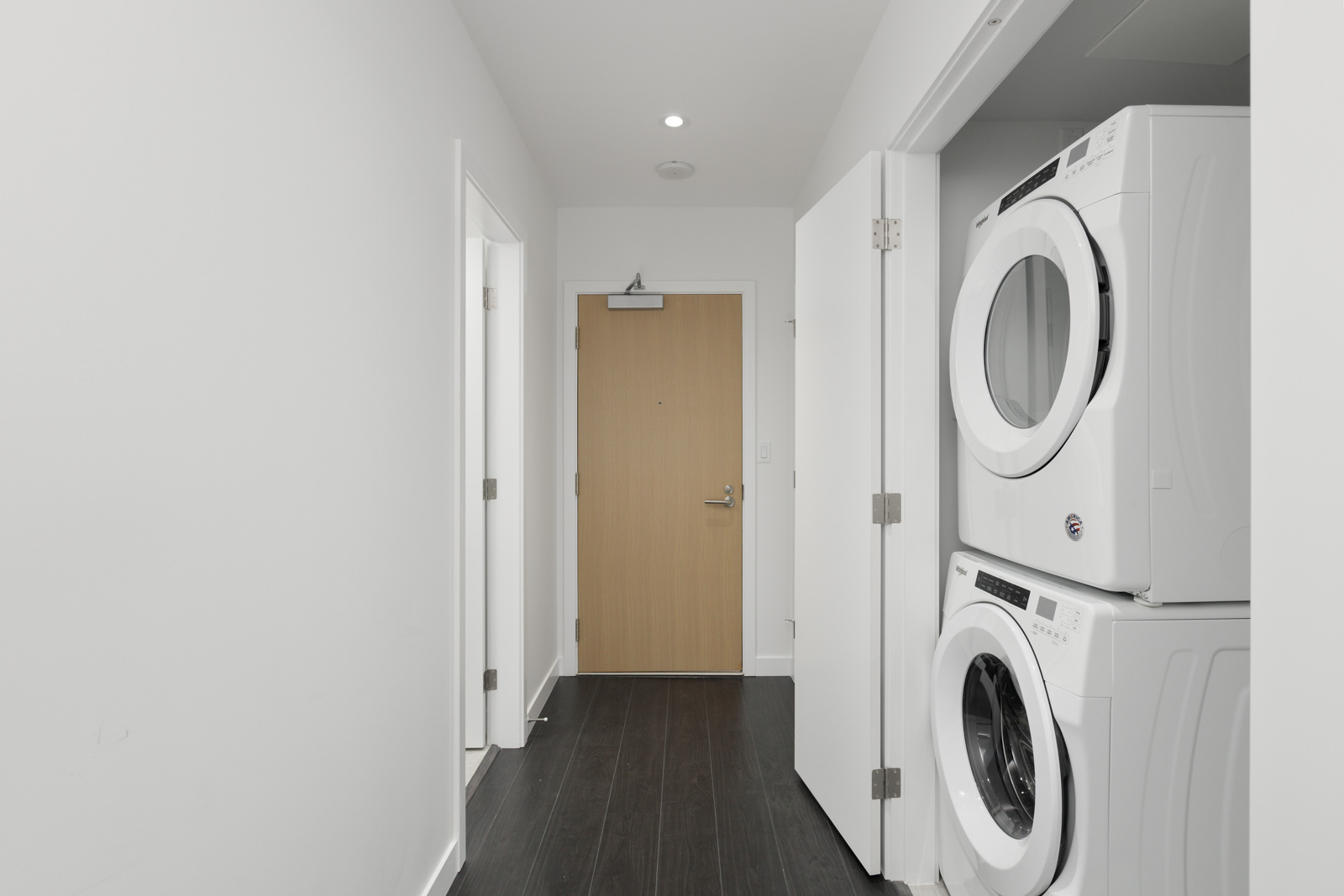A hallway with dark flooring, white walls, a light wood door at the end, and a stacked washer and dryer on the right.