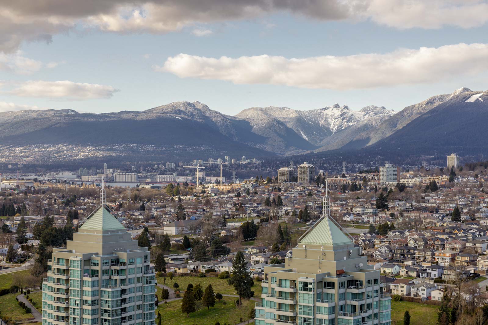 A cityscape with residential buildings in the foreground, a suburban area in the middle ground, and mountains with patches of snow in the background under a partly cloudy sky.