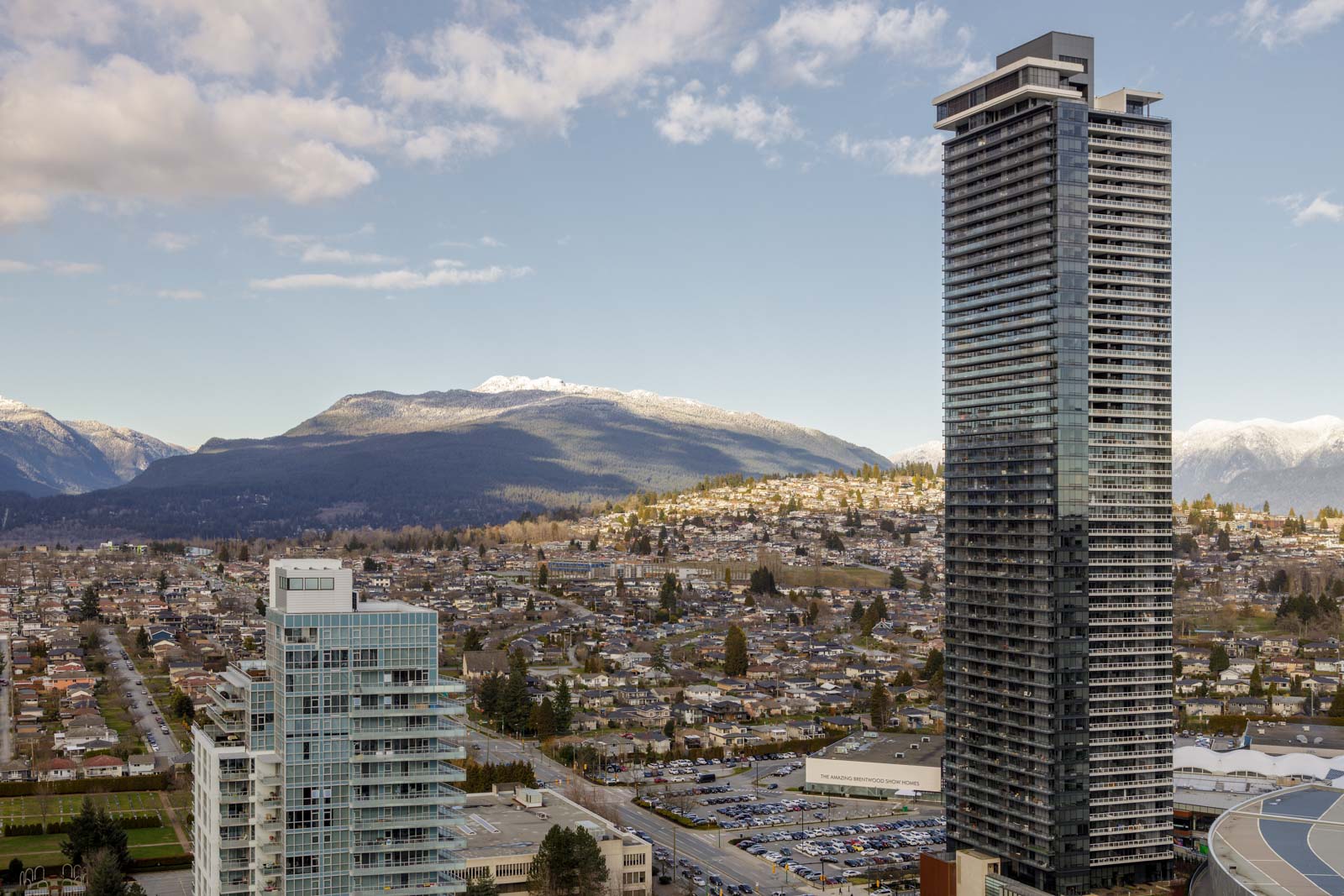 Cityscape with tall modern buildings in the foreground, residential houses, and mountains in the background under a partly cloudy sky.