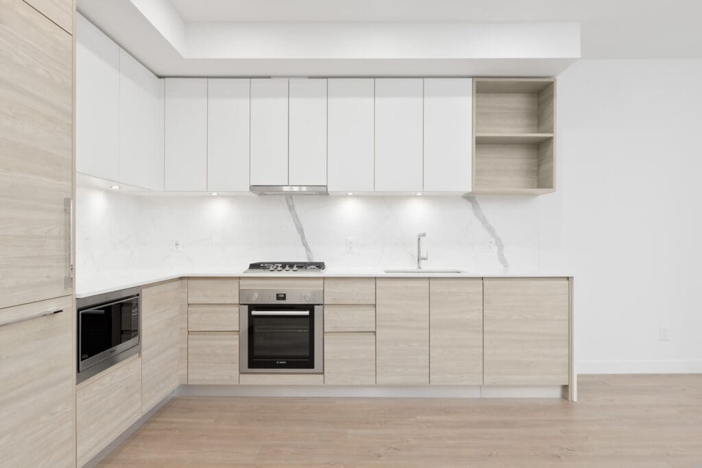Modern kitchen with light wood cabinets, white upper cabinets, marble backsplash, built-in oven, stovetop, microwave, and stainless steel sink.
