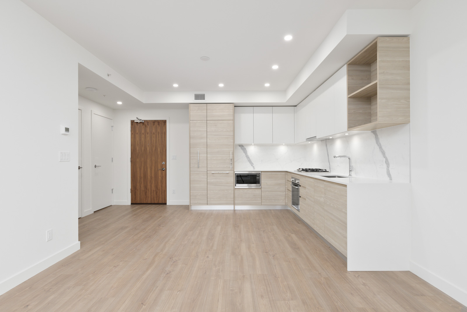 Modern, unfurnished apartment kitchen with light wood floors, white cabinets, built-in appliances, and recessed lighting. A wooden entry door and white walls are visible.