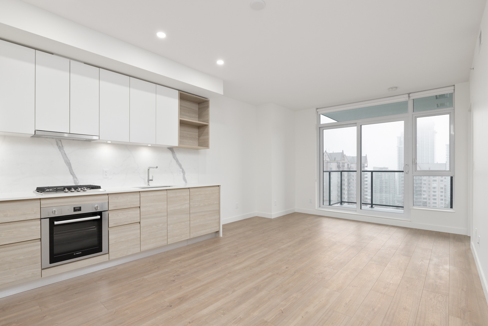 Modern, unfurnished apartment kitchen with light wood floors, built-in oven, minimalist white cabinets, and large window leading to a balcony with city views.