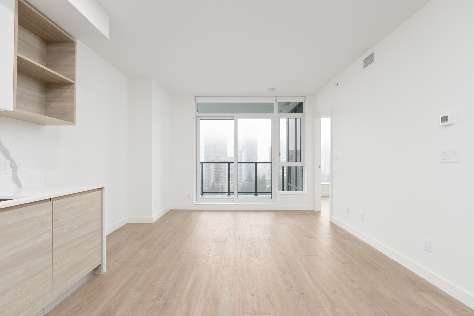 Unfurnished modern apartment with light wood floors, white walls, built-in cabinets, and large windows leading to a balcony overlooking city buildings.